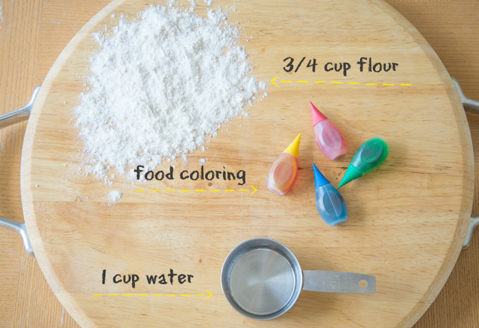 A circular wooden board with flour, food coloring, and a metal cup with water in it and the labels by it.