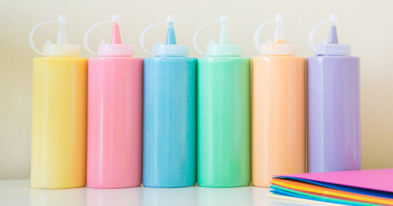 Six squeeze bottles with homemade paint in yellow, pink, blue, green, orange, and purple lined up with a stack of colored paper next to it.