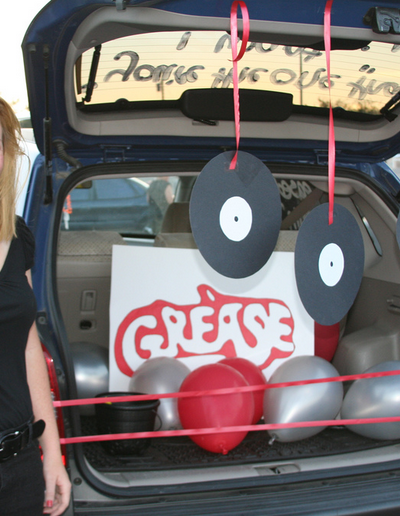 Person standing next to a trunk decorated with paper albums, balloons, and a sign that reads "Grease."