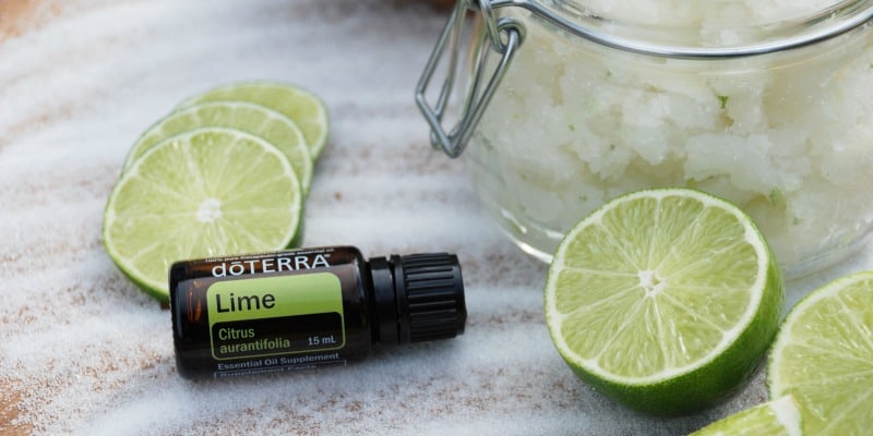 Jar of coconut lime sugar scrub with limes and a bottle of doTerra lime essential oil.