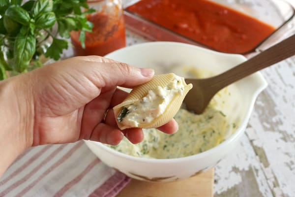 A white bowl with cheese filling and a hand filling a pasta shell with herbs and tomato sauce behind it.