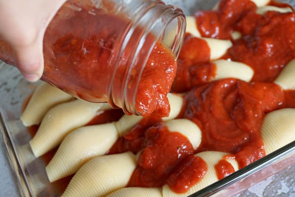 A glass pan with uncooked pasta shells with a hand pouring a glass jar with tomato sauce on it.
