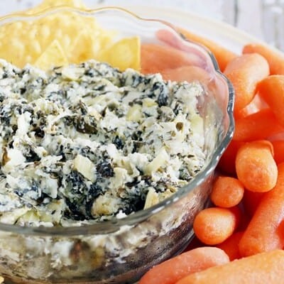 Bowl of spinach artichoke dip surrounded by chips and carrots.