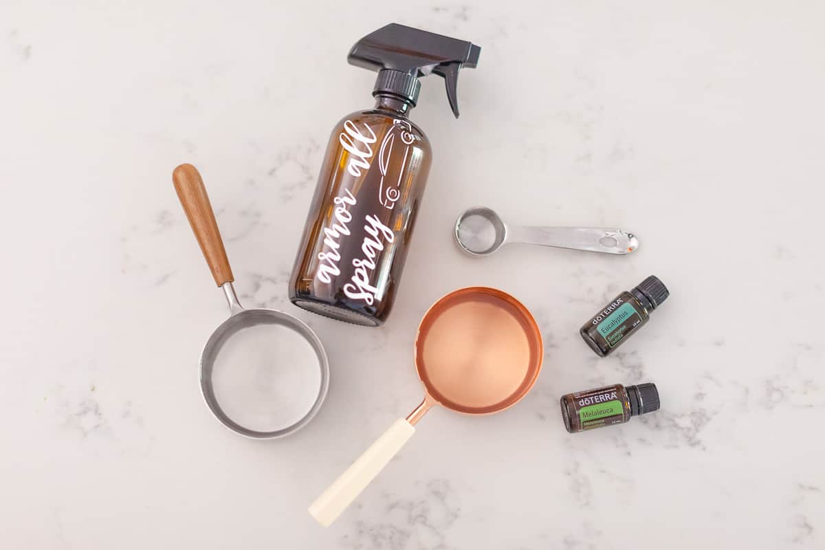 There is a hand holding a silver measuring cup with a wooden handle pouring clear liquid to a white funnel, a silver measuring spoon, a copper measuring cup with a white handle, a Eucalyptus doterra essential oil bottle, a Melaleuca doterra essential oil bottle and a spray bottle top.
