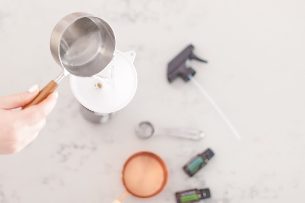 There is a hand holding a silver measuring cup with a wooden handle pouring clear liquid to a white funnel, a silver measuring spoon, a copper measuring cup with a white handle, a Eucalyptus doterra essential oil bottle, a Melaleuca doterra essential oil bottle and a spray bottle top.