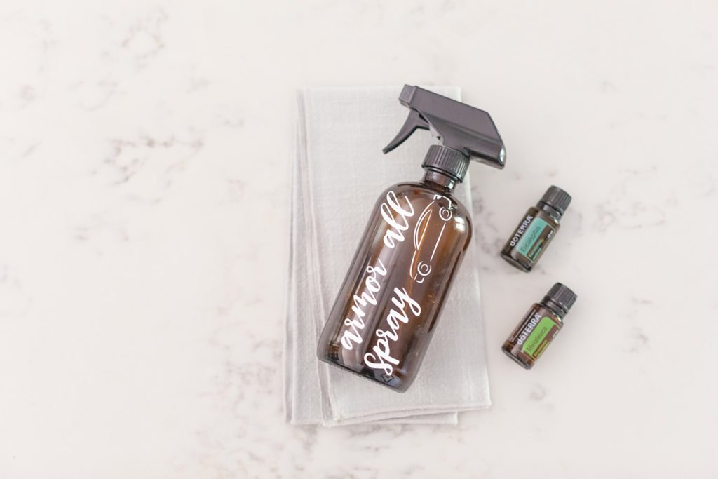 On a white marble background there is a brown spray bottle with the words "armor all spray" in white writing with an image of a car on the bottle on a cloth with a Eucalyptus doterra essential oil bottle and a Melaleuca doterra essential oil bottle on the right side of the bottle.