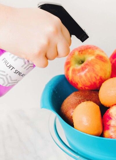 Person using a purple spray bottle labeled "Fruit Spray" on a bowl of fruit.