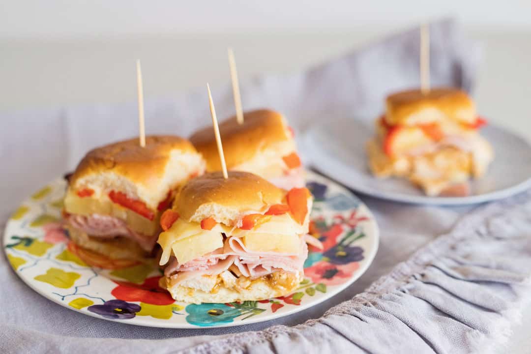 A flowered plate with three ham and cheese sliders with a toothpick in it and a plate with one sandwich behind it.