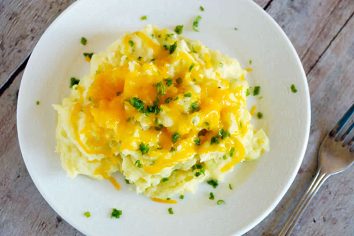 A white bowl of mashed potatoes with cheese and chives on them with a metal fork on the side.