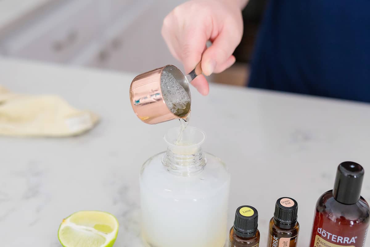  A hand holding a measuring cup pouring soap mixture into the soap canister with bottles and lemon around it.