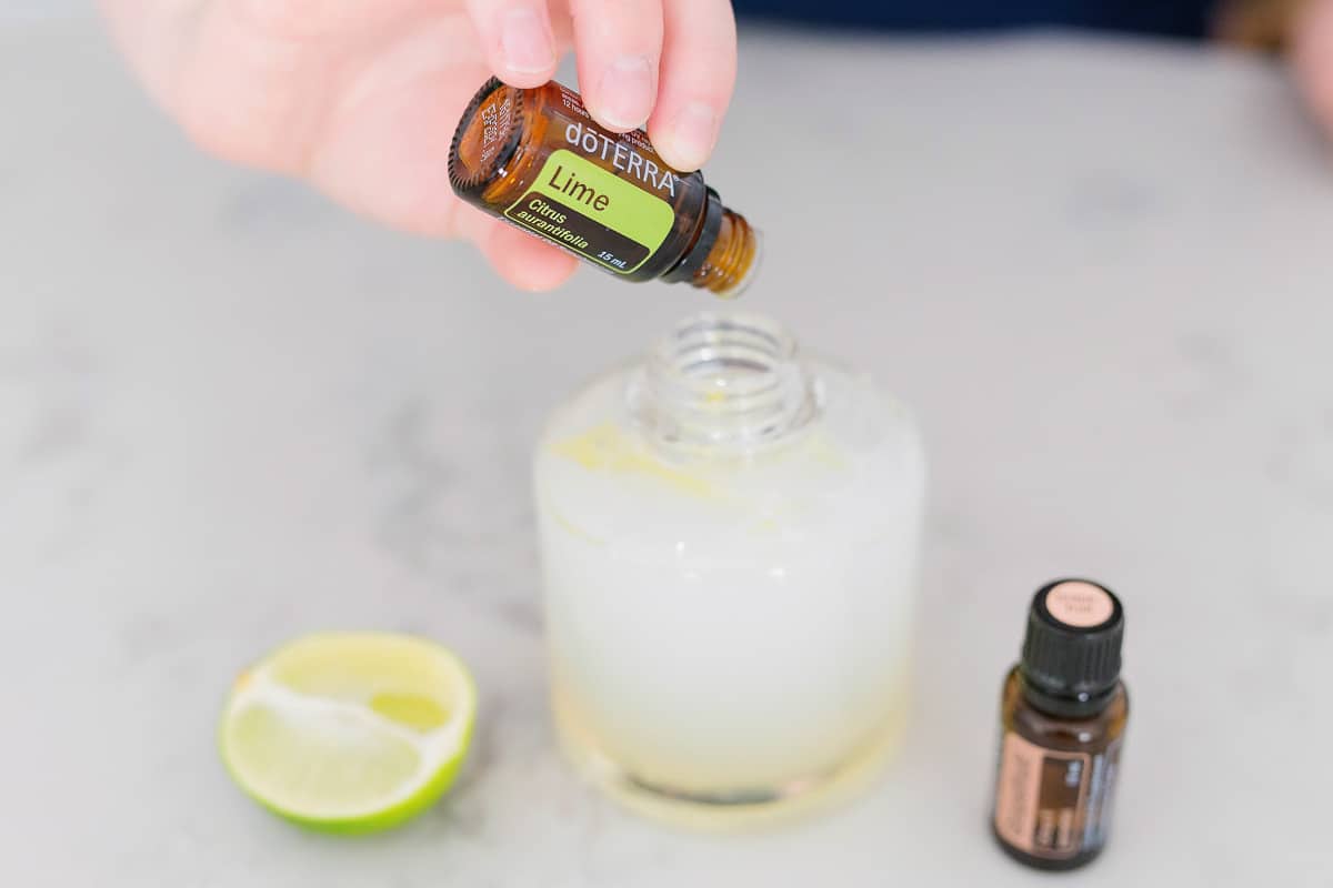 A hand pouring lime essential oil into the soap canister with a cut lime  with another essential oil bottle next to it.