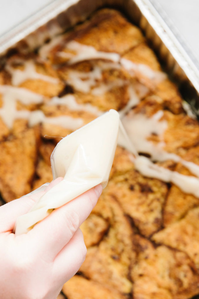 A pan of french toast casserole with a hand pouring out a white bag of icing on it.