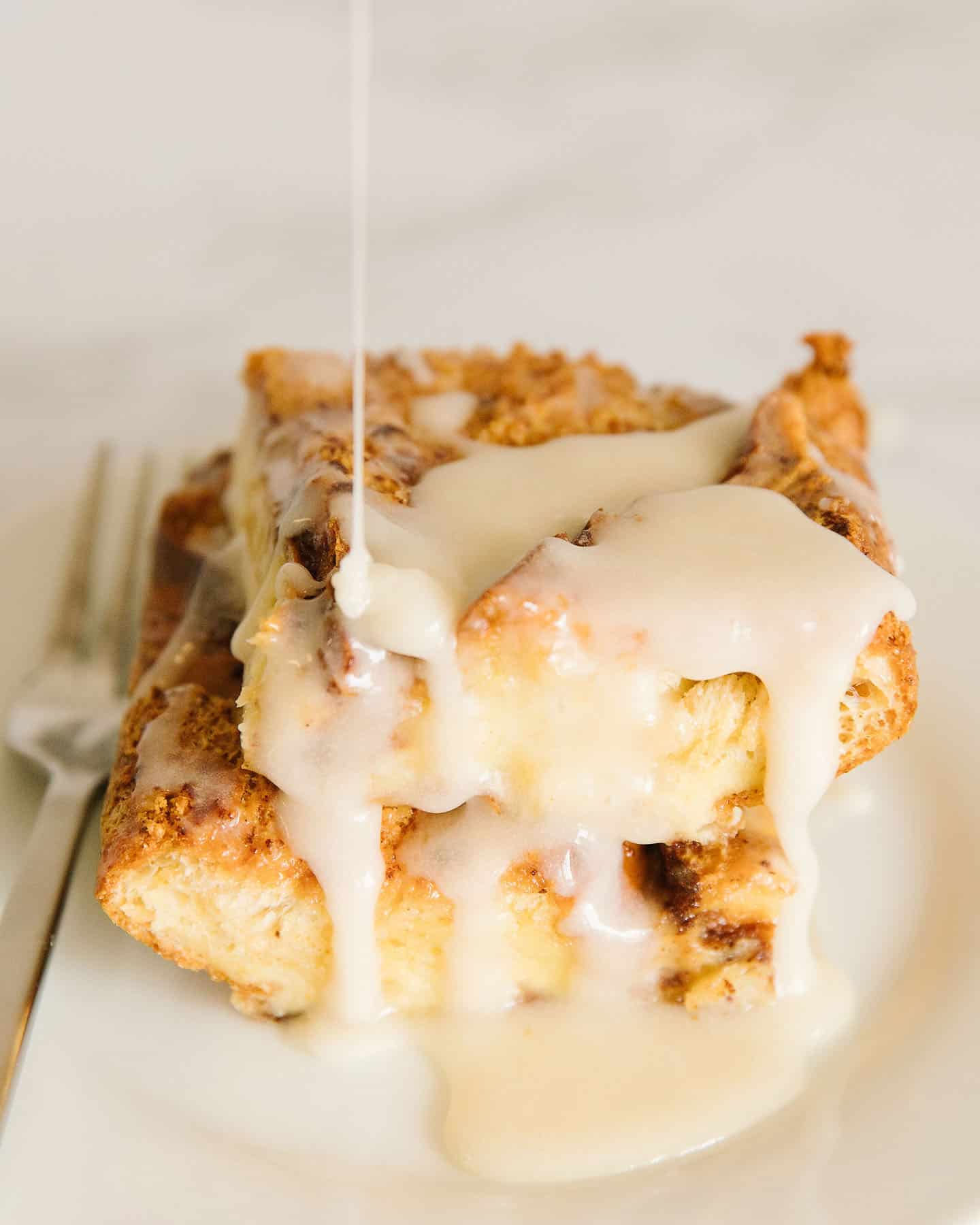 A piece of french toast casserole with icing dripping from the top with a silver fork next to it.