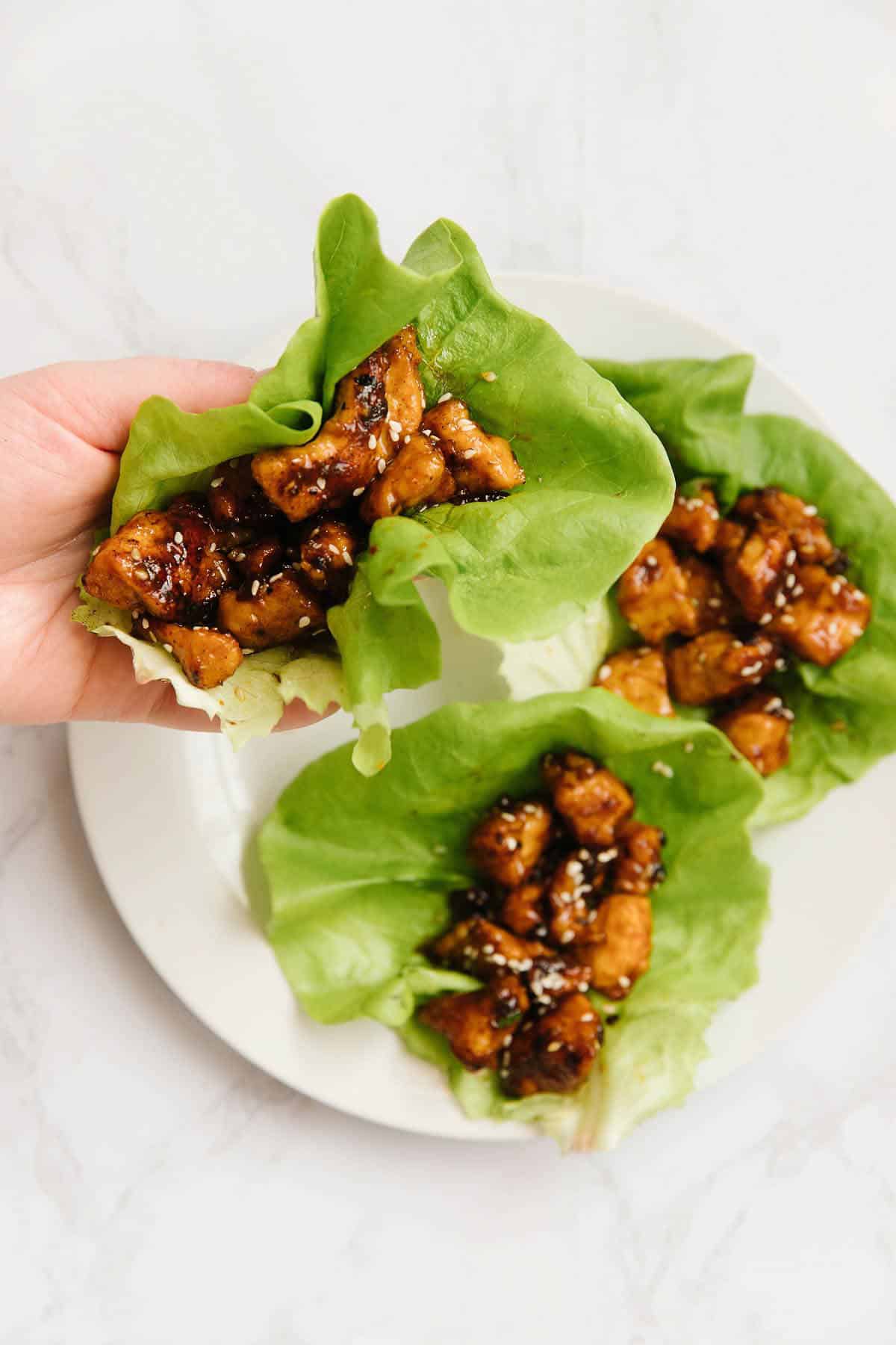 3 lettuce wraps on a white plate - one held in a hand and two remain on a white plate. 