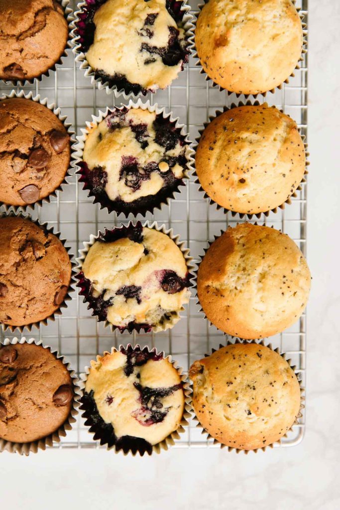 3 different style muffins - poppyseed, blueberry, chocolate - sitting on a wire rack.