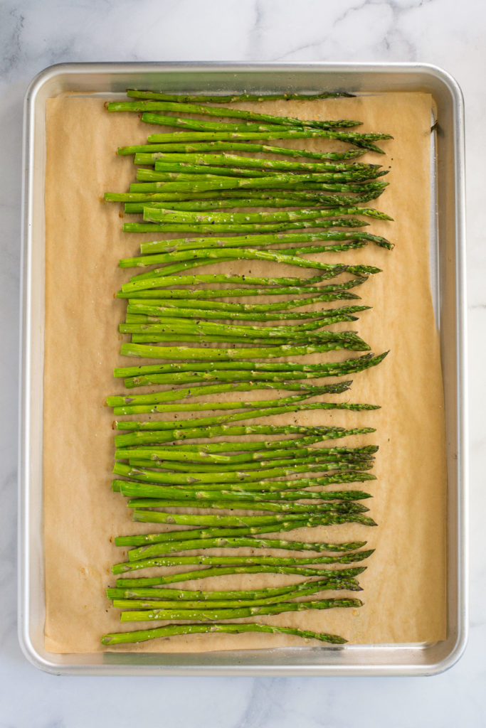 A cookie sheet with parchment paper with asparagus on it sprinkled with salt and pepper.