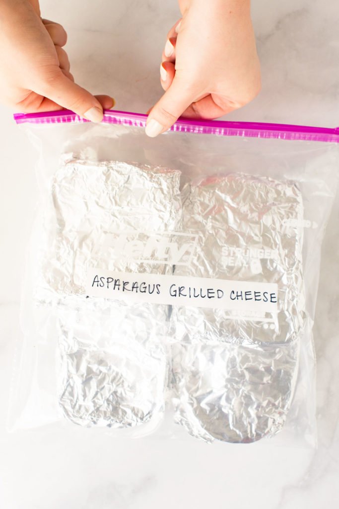 Two hands sealing a Ziploc bag filled with two foil-wrapped sandwiches with the words Asparagus Grilled Cheese.
