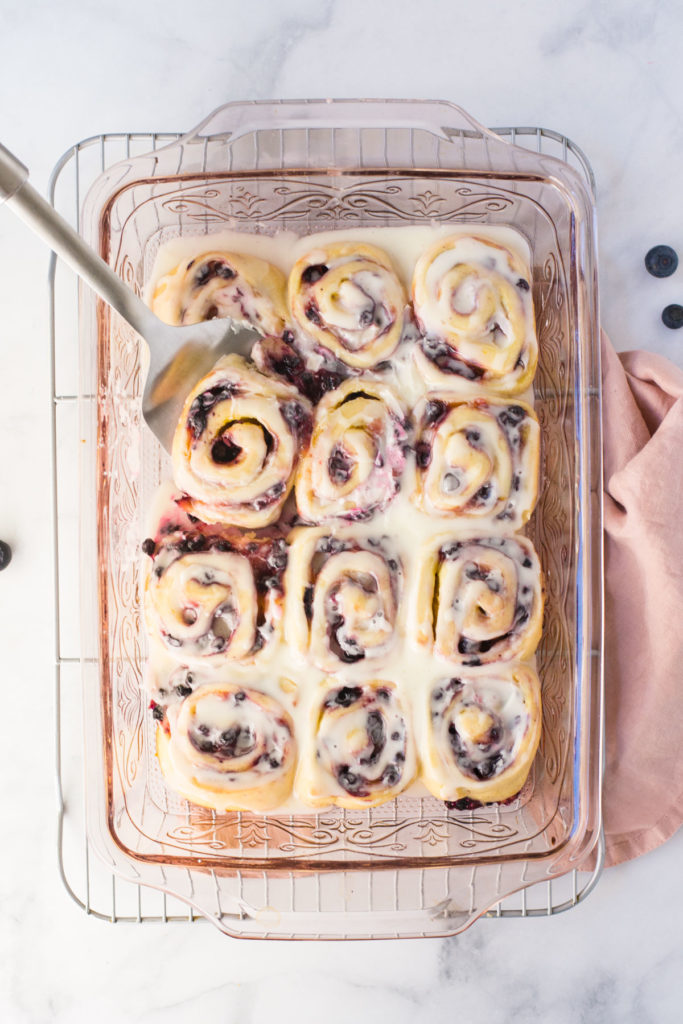 A pink pan holds 12 blueberry lemon sweet rolls topped with white icing. There is a spatula spooning out one roll on the left side and a pink towel underneath the pan with a few blueberries on the side.
