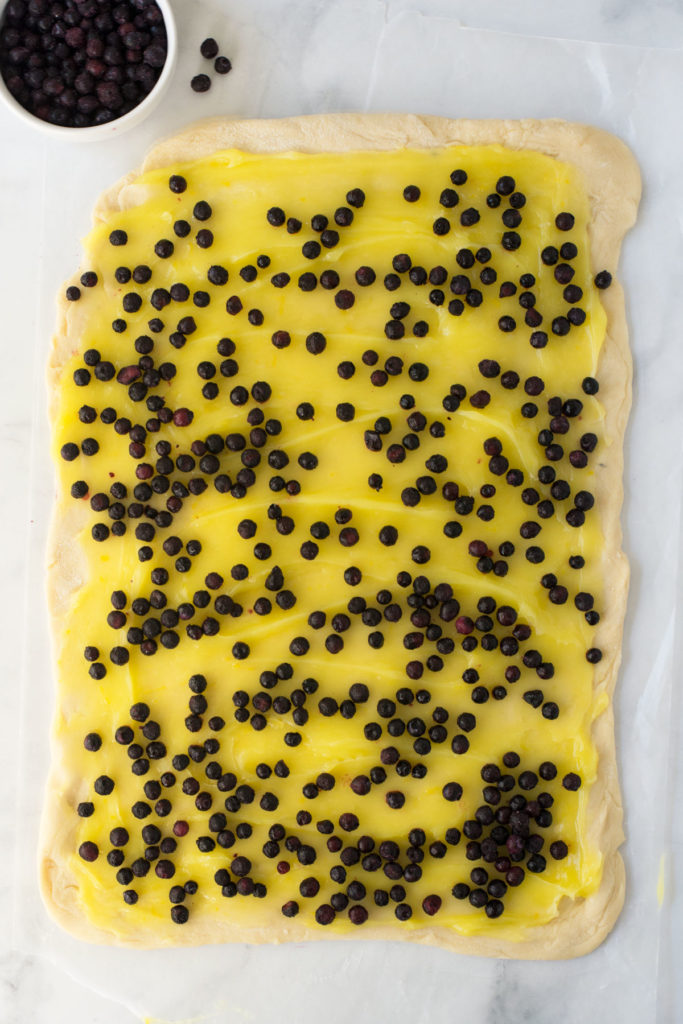 A rectangular piece of rolled out dough with lemon filling and blueberries sprinkled over it. There is a bowl of blueberries in the left corner.