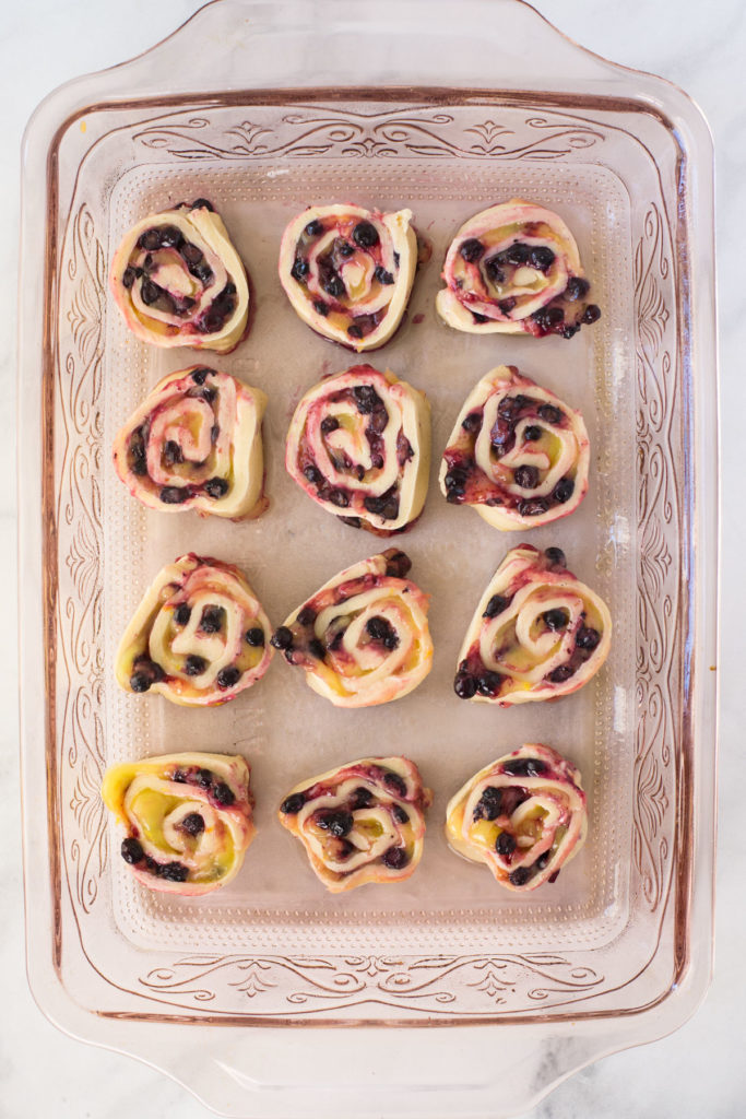 A pink pan holds 12 unbaked dough rolls filled with blueberries and lemon curd.