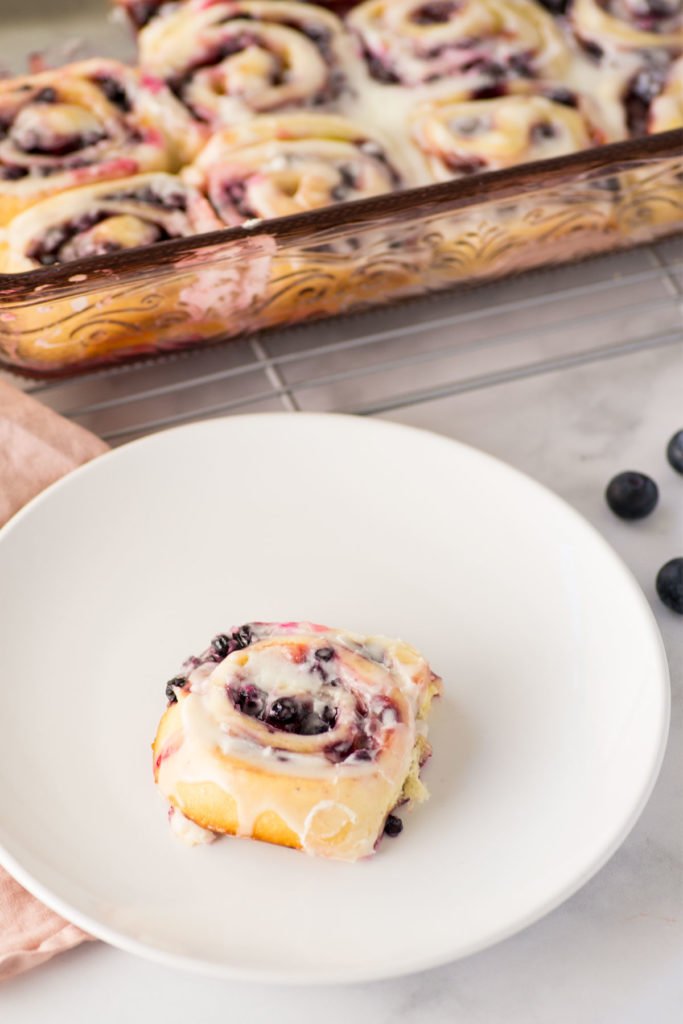 A pink pan holds sweet rolls topped with white icing in the background. A single white circular plate holds one sweet roll. There are a few blueberries on the counter.