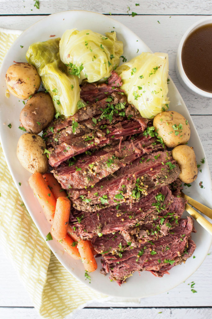 Sliced corned beef covered in spices and herbs with whole yellow potatoes, whole baby carrots and cabbage surround it on a white oval plate. There is a yellow towel under the plate and a white cup of gravy in the right corner.