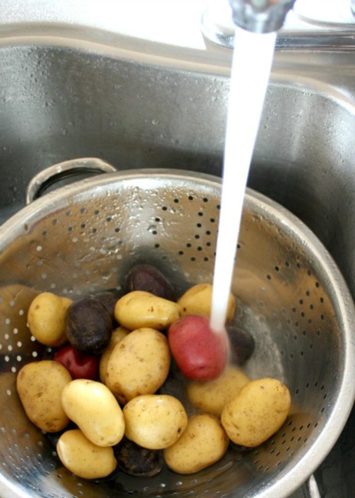 Streaming water in a kitchen sink over a metal strainer filled with whole red, purple and yellow potatoes.
