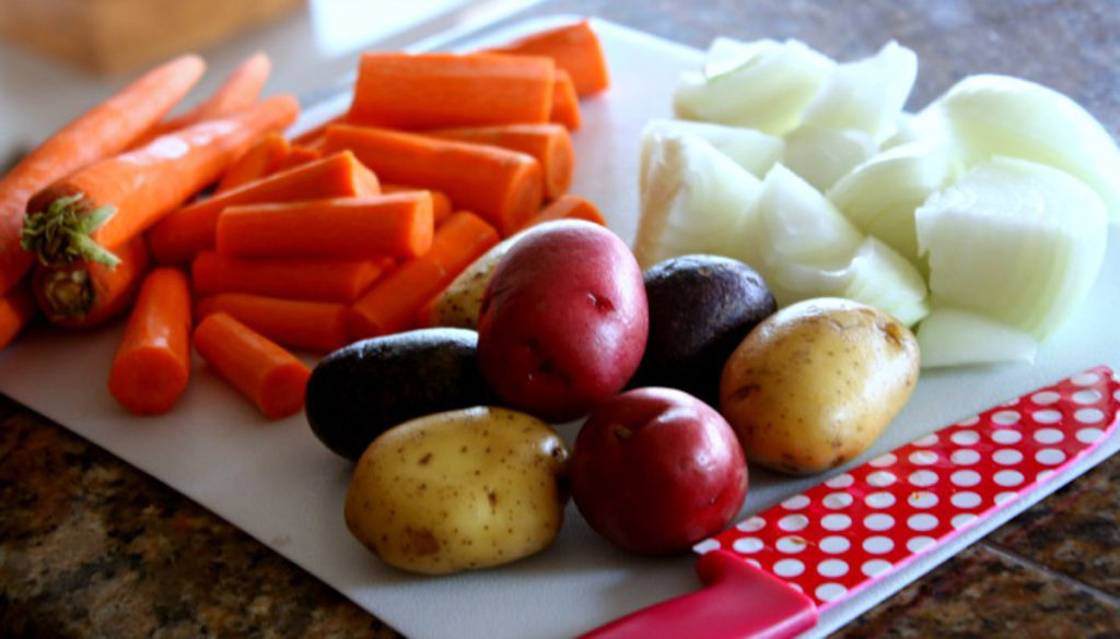 There is a white cutting board with whole carrots, cut carrots, white onion cut up, and whole small red, purple and yellow potatoes.  There is a red and white polka dot knife on the right side.