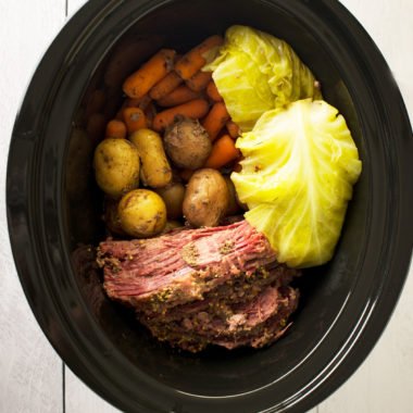 A slow cooker with sliced corned beef with spices. On the side are whole yellow potatoes, whole carrots and pieces of cabbage.
