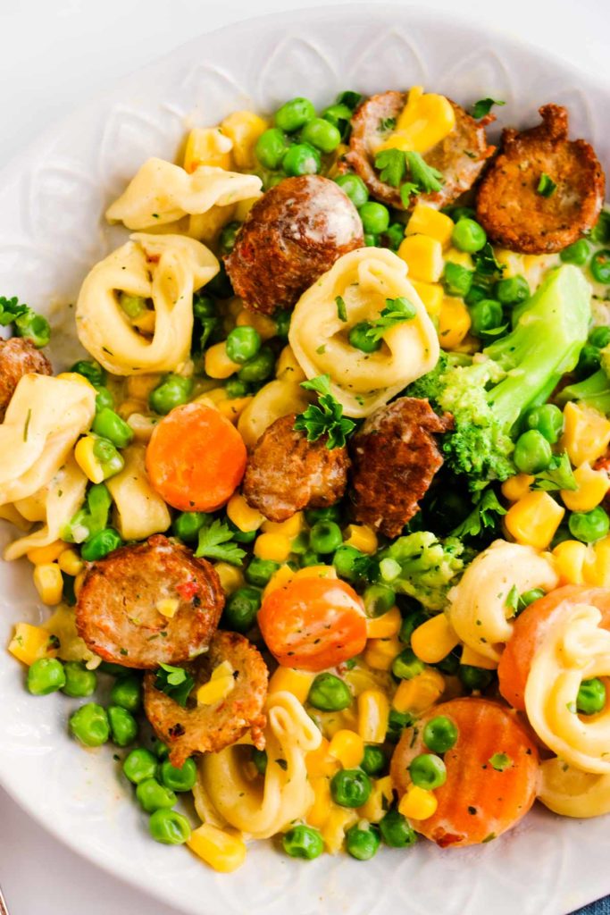 In a big, white bowl holds bite-size pieces of chicken sausage, tortellini, broccoli, carrots, peas and corn in a creamy white sauce.