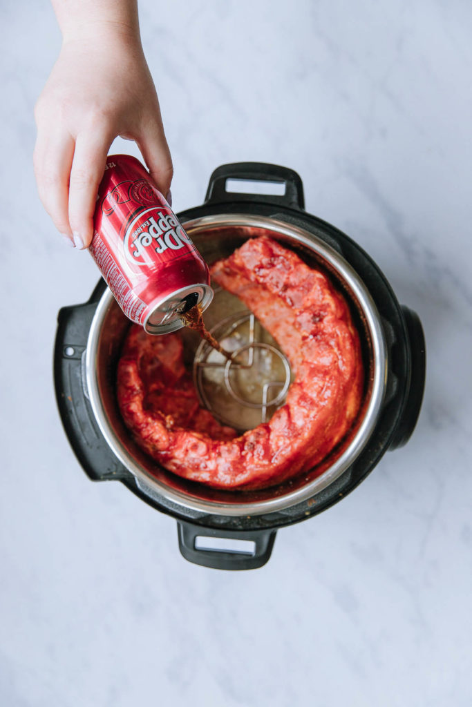 An Instant Pot with uncooked ribs wrapped around the outside with a hand pouring a can of Dr. Pepper inside the middle of it.