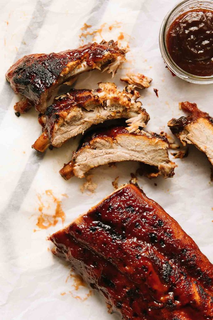 3 ribs cut apart and laid together in a line with a side of bbq sauce in a glass bowl on the top right side. There is also an uncut portion of ribs laying at the bottom.