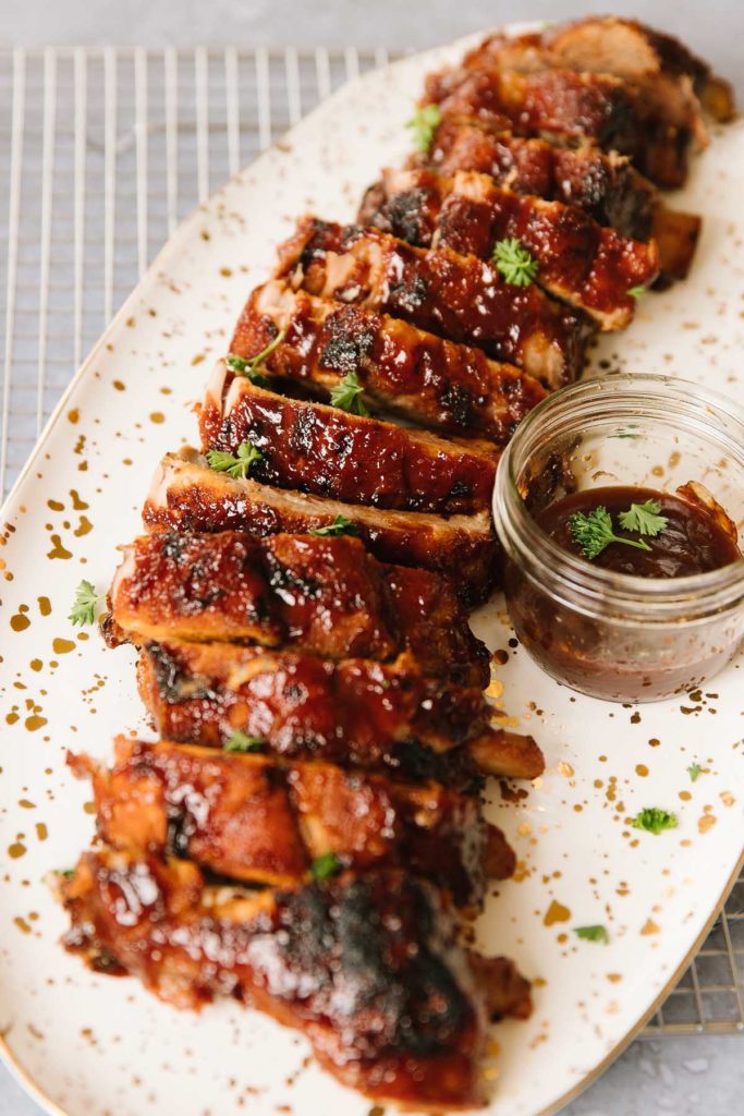 Ribs cut apart and laid together in a line smothered in bbq sauce sprinkled with herbs on a white oval plate with a side of bbq sauce in a glass bowl on the right side.