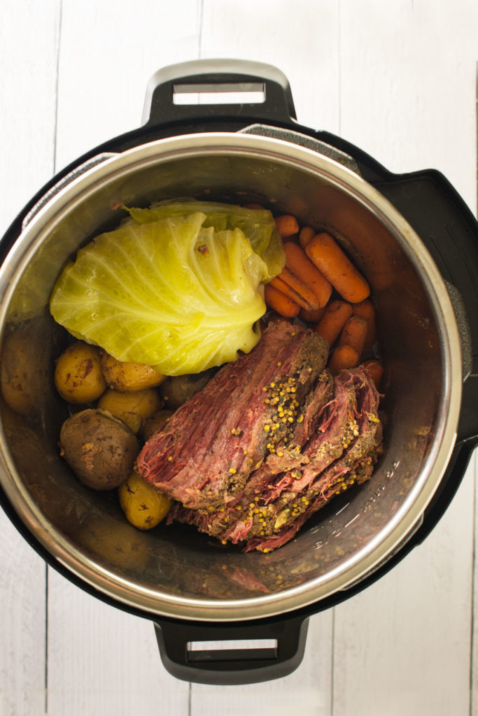 An instant pot with a sliced corned beef covered in spices and whole small red, purple and yellow potatoes, whole baby carrots and cabbage surround it.