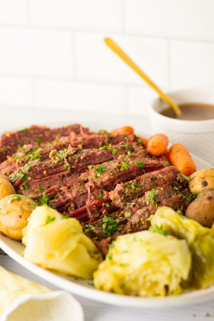Sliced corned beef covered in spices and herbs with whole yellow potatoes, whole baby carrots and cabbage surround it on a white oval plate. There is a peek of a yellow towel under the left corner and a white cup of gravy with a yellow spoon in it in the right corner.