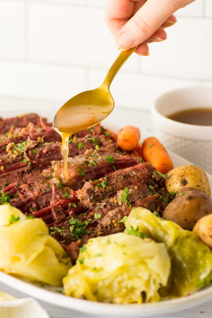 Sliced corned beef covered in spices and herbs with whole yellow potatoes, whole baby carrots and cabbage surround it on a white oval plate. A golden spoon is spooning gravy over the corned beef slices. There is a peek of a yellow towel under the left corner and a white cup of gravy in the right corner.