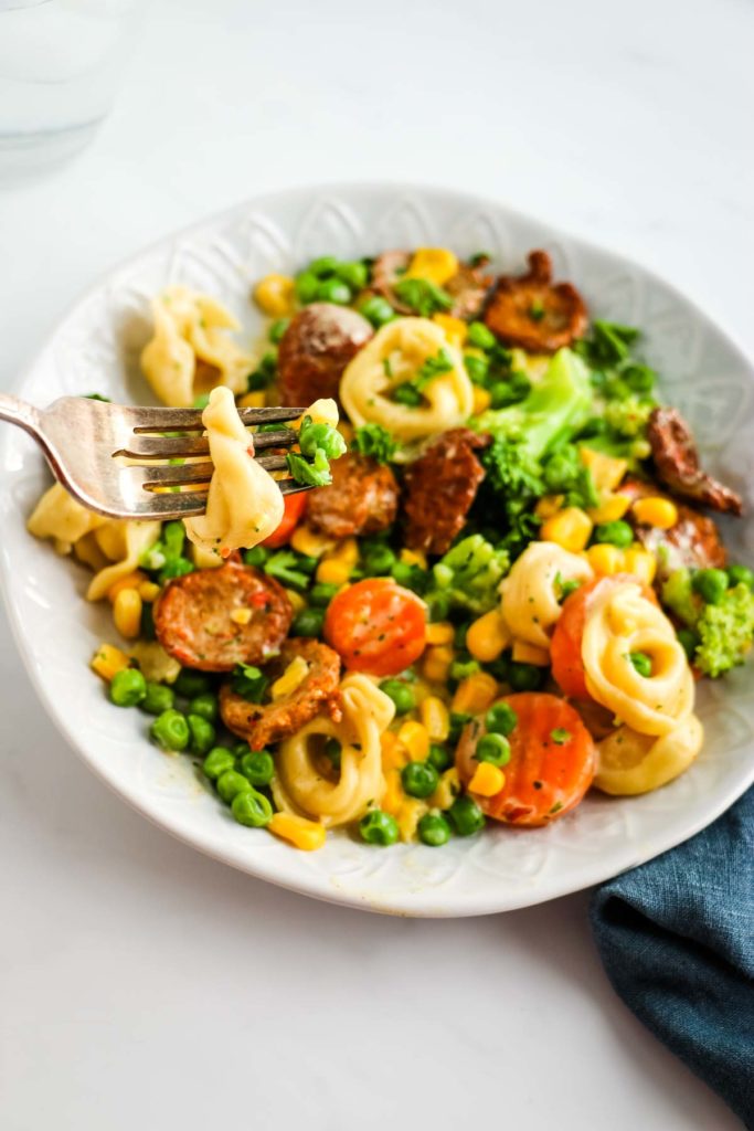 There is a white decorative bowl with pieces of chicken sausage, tortellini, corn, peas, broccoli, carrots.  Above the bowl is a fork with tortellini and a small piece of broccoli. Underneath the bowl is a blue denim napkin under the right lower side of the bowl.