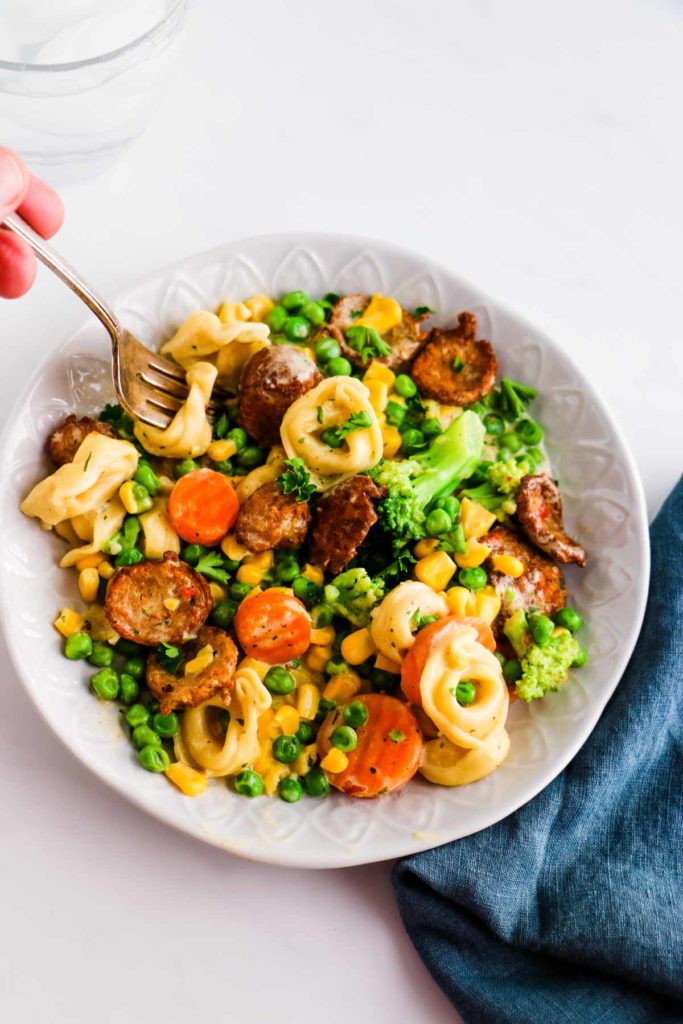 There is a white decorative bowl with pieces of chicken sausage, tortellini, corn, peas, broccoli, carrots.  In the bowl is a fork spearing a tortellini and veggies. Underneath the bowl is a blue denim napkin under the right lower side of the bowl.