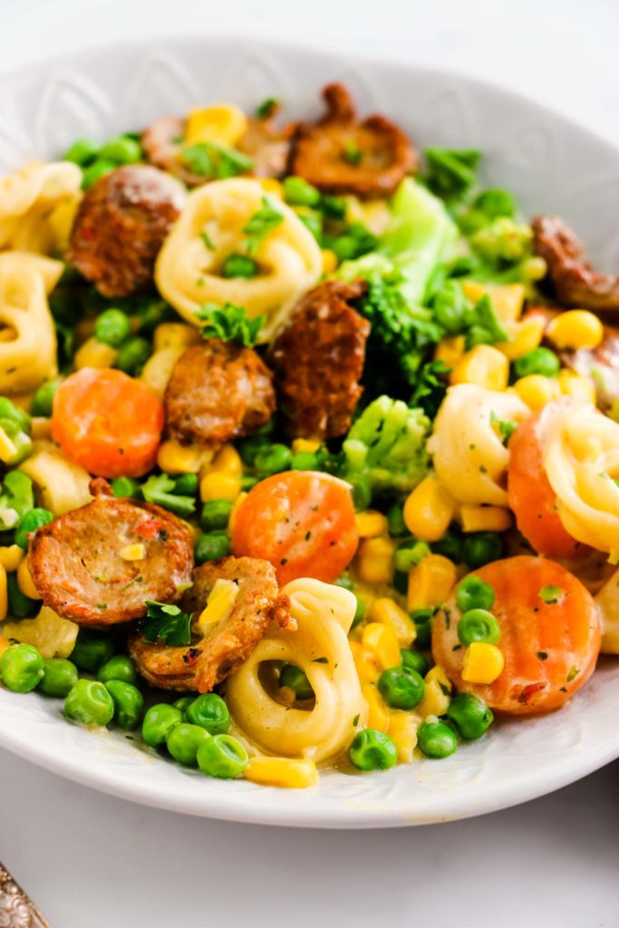 There is a white decorative bowl with pieces of chicken sausage, tortellini, corn, peas, broccoli, carrots.  The bottom of the left side is a little piece of a the metal fork.
