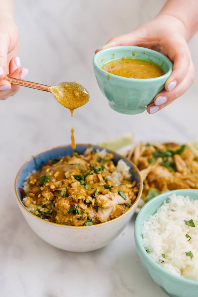 There are two bowls in this picture.  The first bowl has peanut chicken satay with green onions on it. The other bowl has rice with green onions on it. There is a plate in the back right corner with chicken satay blurred in the background. Two hands are holding a small green bowl with peanut sauce and a spoon dropping sauce on the peanut satay.