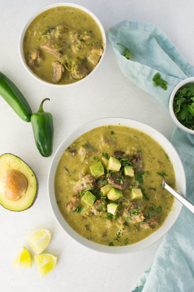 Two bowls of pork green chili verde. One bowl has chopped avocado on it with cilantro. There is a small bowl of cilantro on the side with half an avocado, jalapenos, and lime scattered around.