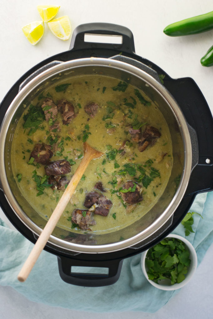 An instant pot filled with pork chili verde with a wooden spoon in it. There are limes, jalapenos, and a small bowl of cilantro under a blue towel.