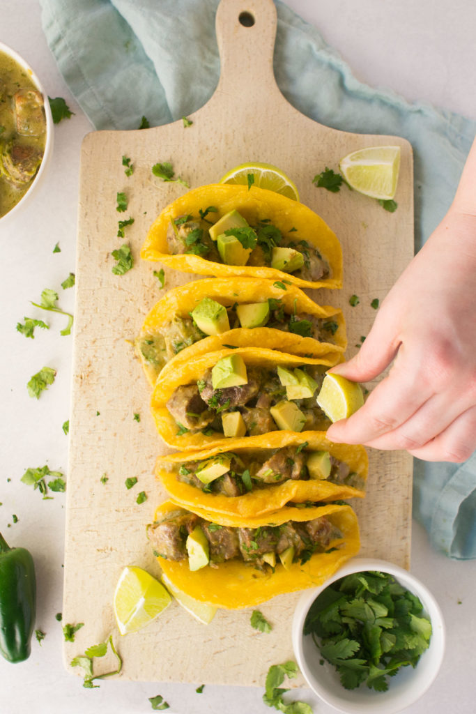 A cutting board filled with a line of 5 street tacos stuffed with pork chili verde. There is a hand squeezing fresh lime over the tacos with a small bowl of cilantro, jalapeno and pork verde in a bowl on the side.