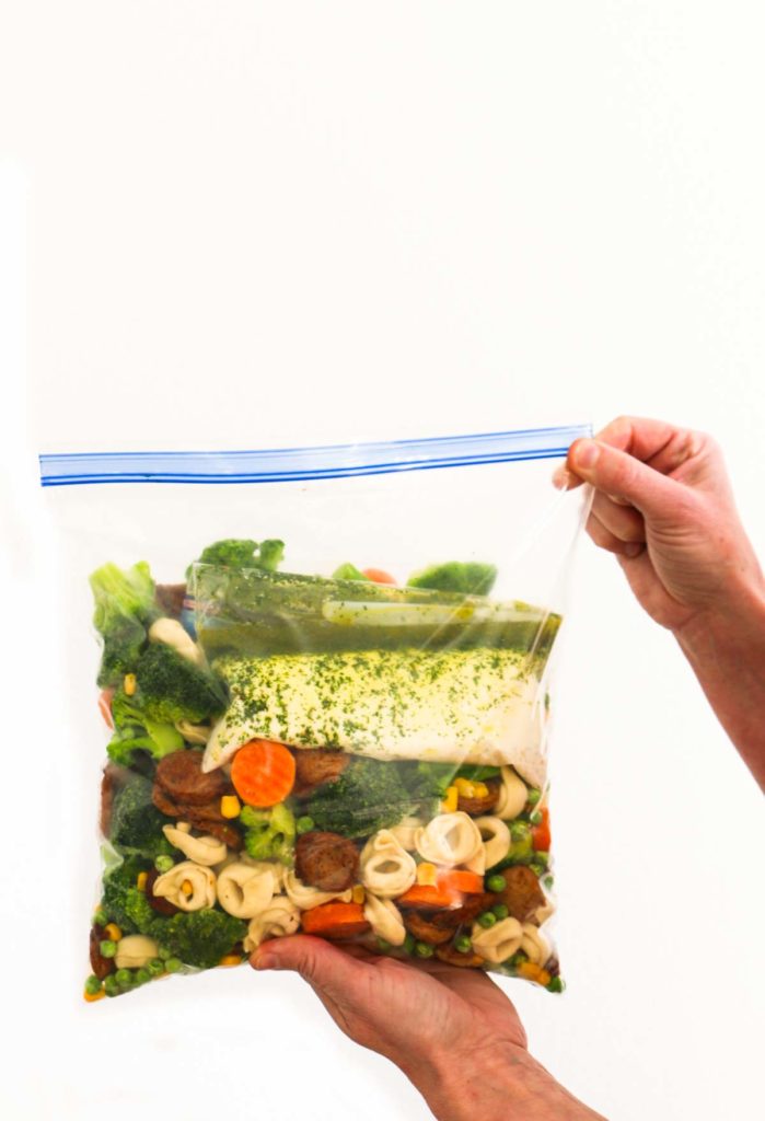 A gallon-size ziploc bag  filled with pieces of chicken sausage, tortellini, corn, peas, broccoli, carrots.  There is a smaller ziploc bag inside with a creamy white sauce also zipped in the bag.  There are two hands holding the bag at the bottom and on the right side of the big bag.