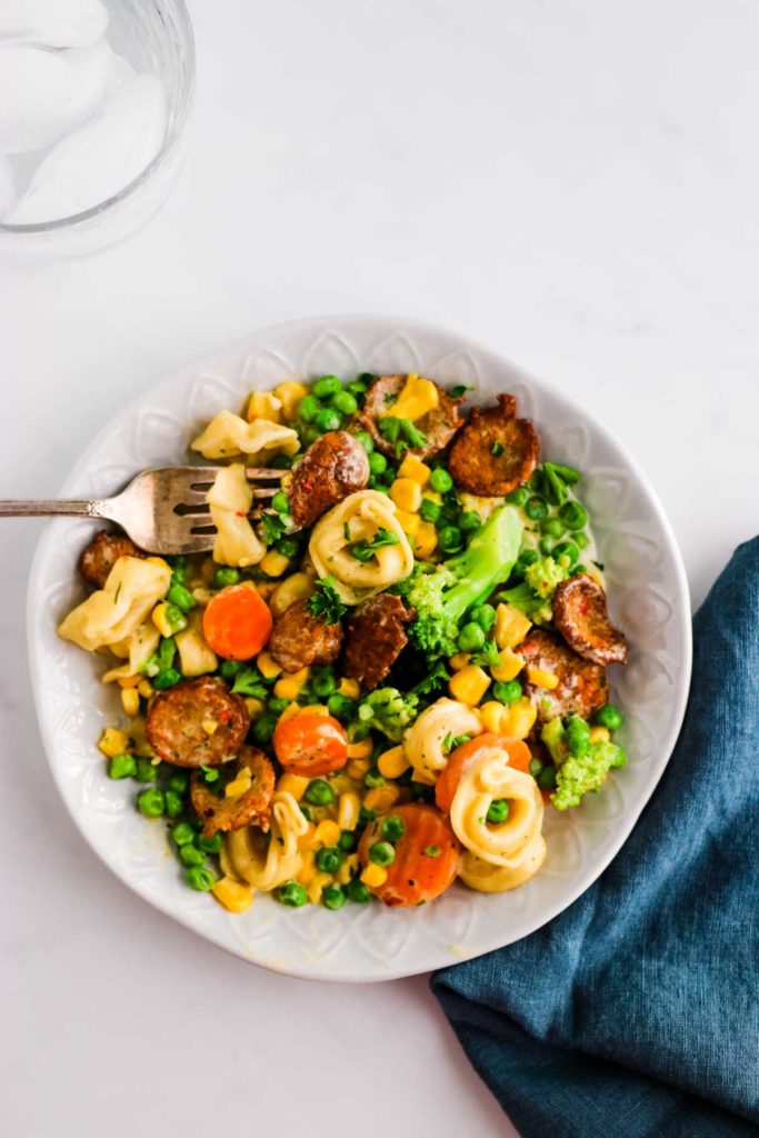 There is a white decorative bowl with pieces of chicken sausage, tortellini, corn, peas, broccoli, carrots.  In the bowl is a fork spearing a tortellini, veggies and a piece of chicken sausage. Underneath the bowl is a blue denim napkin under the right lower side of the bowl.