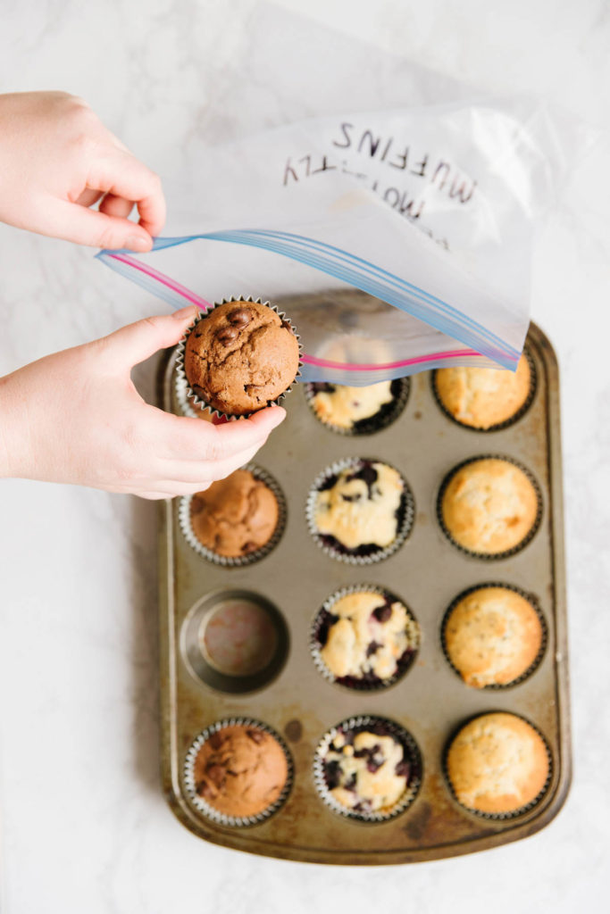 A muffin tin with eleven muffins - chocolate, poppyseed, blueberry - in it. Two hands are holding a gallon size ziploc bag with one muffin being put in it.