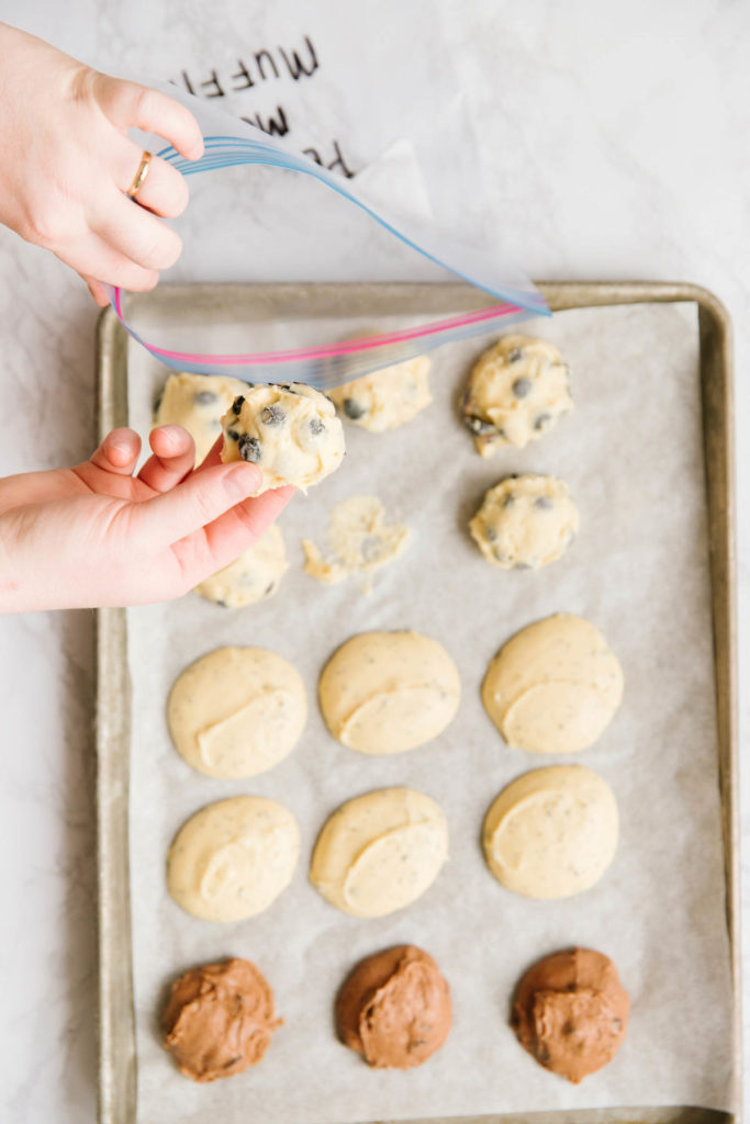 A cookie sheet with wax paper with uncooked frozen muffins of chocolate, poppyseed, and blueberry. There is a gallon size ziploc bag open with two hands putting one of the muffins in it.