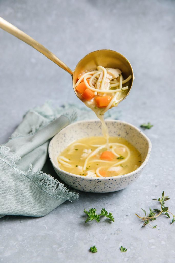 A bowl of chicken noodle soup with a gold ladle spooning more soup in it with a light blue towel on the left side with herbs next to it.