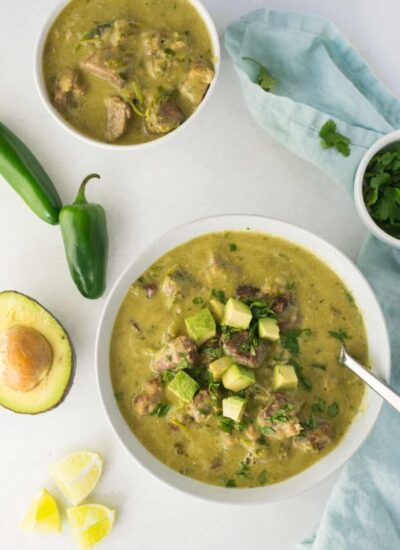 Bowl of pork chili verde topped with pieces of avocado on a counter with various ingredients.