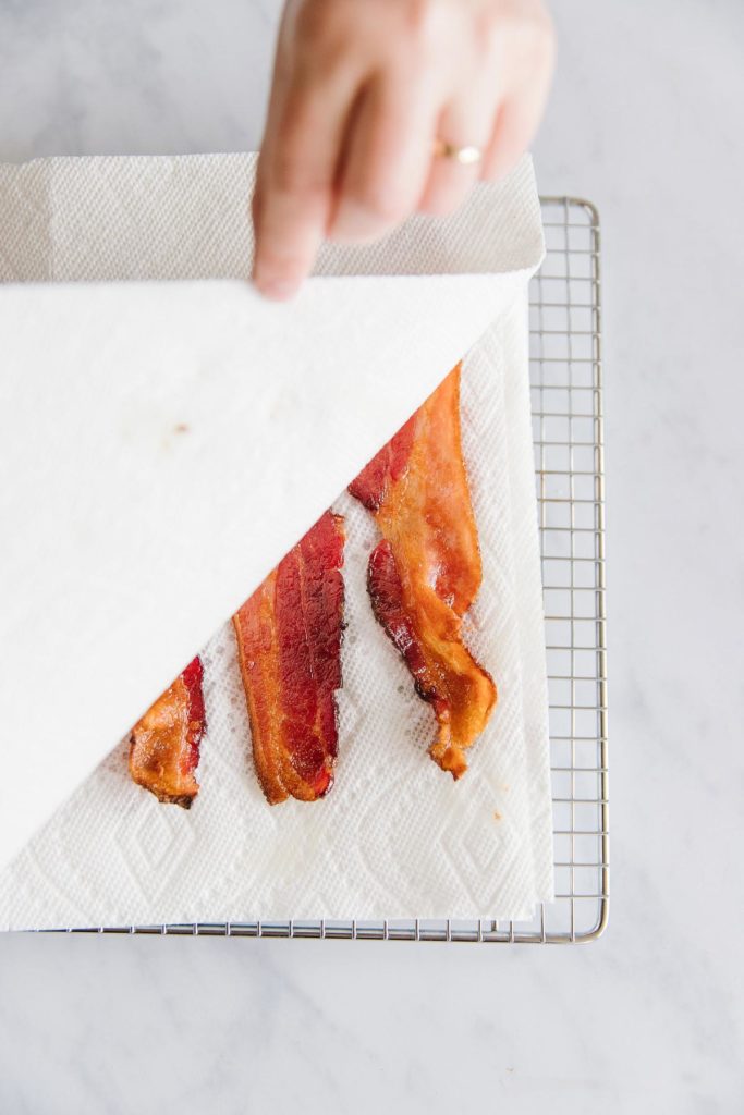 3 strips of bacon with a paper towel above and below sitting on a rack with a hand pulling back half of the paper towel.